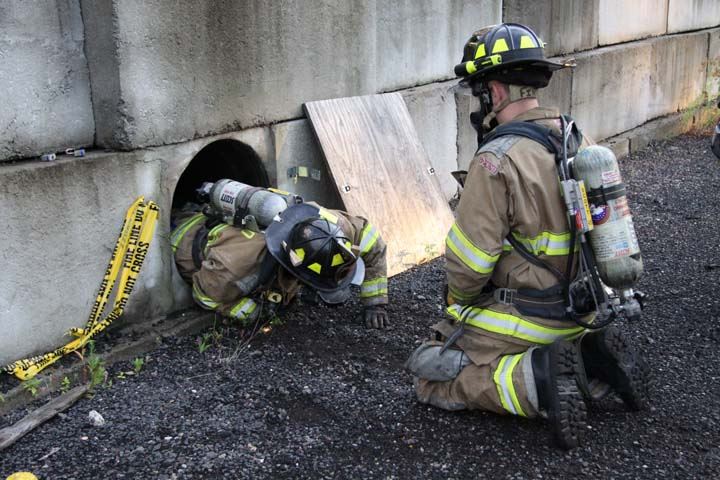 Firefighters in gear entering pipe set in concrete wall